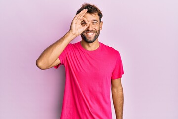 Handsome man with beard wearing casual pink tshirt over pink background smiling happy doing ok sign with hand on eye looking through fingers