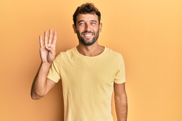 Handsome man with beard wearing casual yellow tshirt over yellow background showing and pointing up with fingers number four while smiling confident and happy.