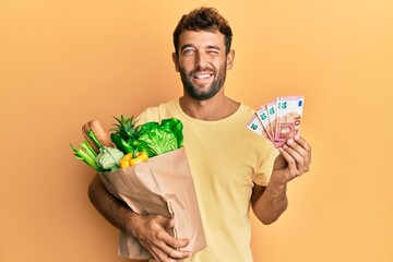 Handsome man with beard holding groceries and 10 euros banknotes winking looking at the camera with...