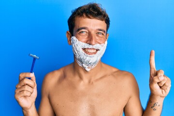 Handsome man saving beard with shave foam over face holding razor smiling with an idea or question pointing finger with happy face, number one