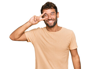 Handsome young man with beard wearing casual tshirt doing peace symbol with fingers over face,...