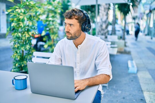 Handsome man with beard wearing casual white shirt on a sunny day working using laptop and wearing headphones at cafeteria