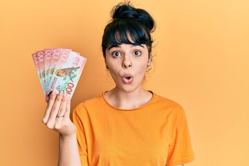 Young hispanic girl holding 100 new zealand dollars banknote scared and amazed with open mouth for surprise, disbelief face