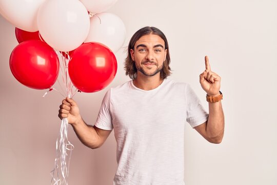 Young handsome man holding balloons smiling with an idea or question pointing finger with happy face, number one