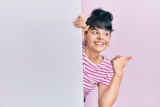 Young hispanic girl holding blank empty banner pointing thumb up to the side smiling happy with open mouth
