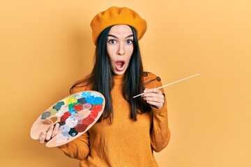 Young brunette woman holding paintbrush and palette wearing beret afraid and shocked with surprise and amazed expression, fear and excited face.