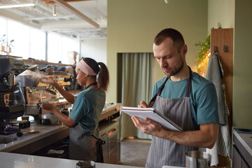 Waist up portrait of male barista wearing apron while working in coffee shop and holding clipboard,...