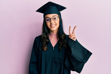 Young hispanic woman wearing graduation cap and ceremony robe showing and pointing up with fingers number two while smiling confident and happy.