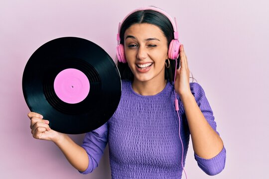 Young hispanic woman using headphones holding vinyl disc winking looking at the camera with sexy expression, cheerful and happy face.