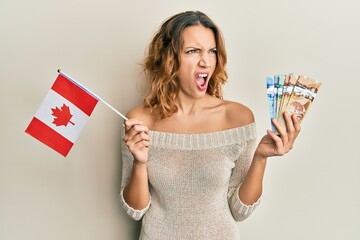 Young caucasian woman holding canada flag and dollars angry and mad screaming frustrated and...