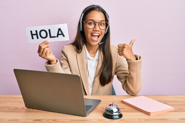Beautiful hispanic woman wearing operator headset holding hola greenting pointing thumb up to the side smiling happy with open mouth © Krakenimages.com
