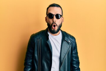 Obraz premium Young man with beard wearing black leather jacket and sunglasses scared and amazed with open mouth for surprise, disbelief face
