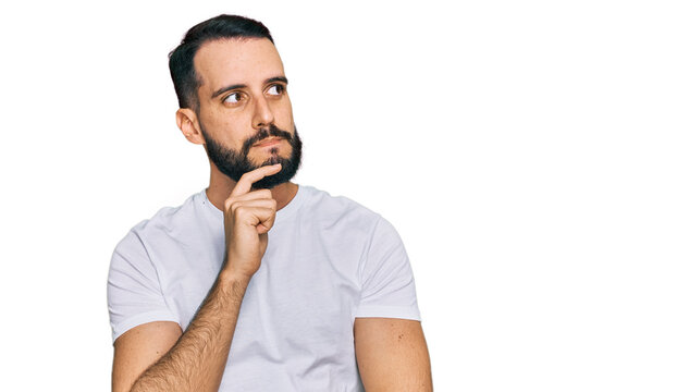 Young man with beard wearing casual white t shirt with hand on chin thinking about question, pensive expression. smiling with thoughtful face. doubt concept.