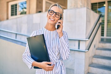 Young blonde businesswoman holding folder and talking on the smartphone at the city.
