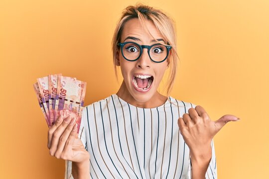 Beautiful blonde woman holding south african 50 rand banknotes pointing thumb up to the side smiling happy with open mouth