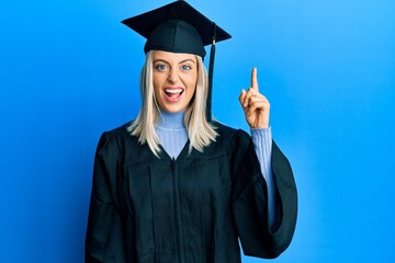 Beautiful blonde woman wearing graduation cap and ceremony robe pointing finger up with successful...