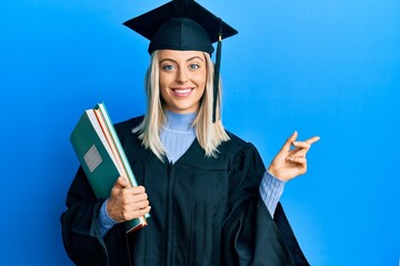 Beautiful blonde woman wearing graduation cap and ceremony robe holding books smiling happy pointing with hand and finger to the side