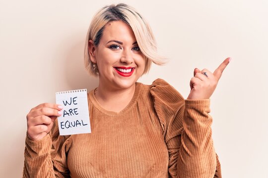 Beautiful blonde plus size woman asking for equality holding paper with we are equal message smiling happy pointing with hand and finger to the side