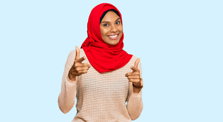 Young african american woman wearing traditional islamic hijab scarf pointing fingers to camera with happy and funny face. good energy and vibes.