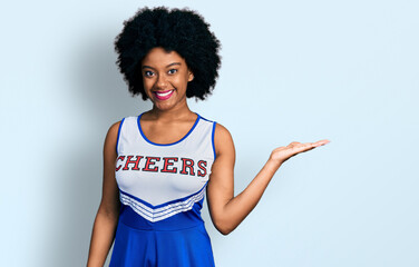 Young african american woman wearing cheerleader uniform smiling cheerful presenting and pointing with palm of hand looking at the camera.