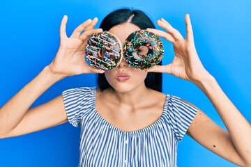 Beautiful young woman holding tasty colorful doughnuts on eyes skeptic and nervous, frowning upset because of problem. negative person.