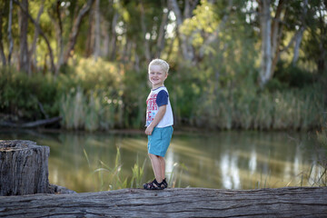 Little blonde caucasian boy enjoying outdoor adventure by the river climbing on old logs