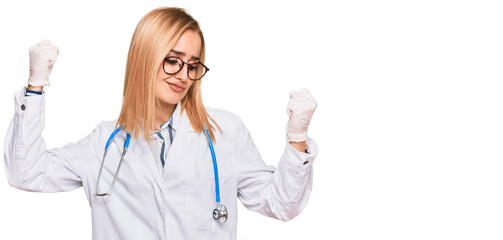 Beautiful caucasian woman wearing doctor uniform and stethoscope showing arms muscles smiling proud. fitness concept.