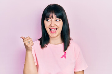 Young hispanic woman wearing pink cancer ribbon on shirt pointing thumb up to the side smiling happy with open mouth