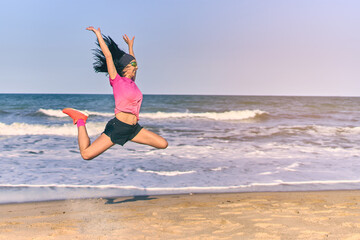 Attractive young adult woman jumping on the beach. Happy girl jumps with arms up and bending the legs.
