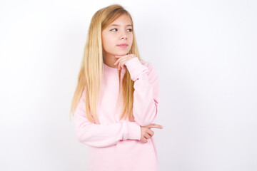 Dreamy beautiful caucasian little girl wearing pink hoodie over white background with pleasant expression, looks sideways, keeps hand under chin, thinks about something pleasant.