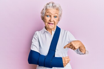 Senior grey-haired woman wearing arm on sling smiling happy pointing with hand and finger