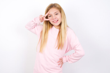 beautiful caucasian little girl wearing pink hoodie over white background making v-sign near eyes. Leisure, coquettish, celebration, and flirt concept.