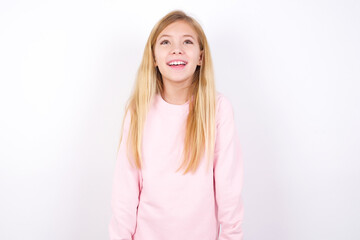 Surprised beautiful caucasian little girl wearing pink hoodie over white background, shrugs shoulders, looking sideways, being happy and excited. Sudden reactions concept.