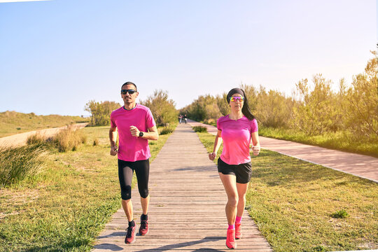 Active couple running during a warm summer day. Wearing pink shirts and short pants. Both are wearing sunglasses. Running towards the camera. Rural landscape.