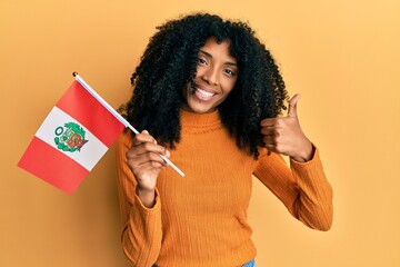 African american woman with afro hair holding peru flag smiling happy and positive, thumb up doing excellent and approval sign