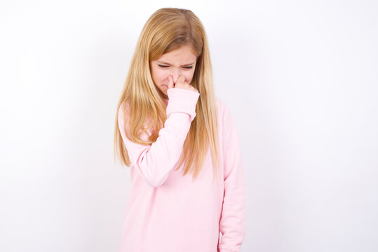 beautiful caucasian little girl wearing pink hoodie over white background, holding his nose because of a bad smell.