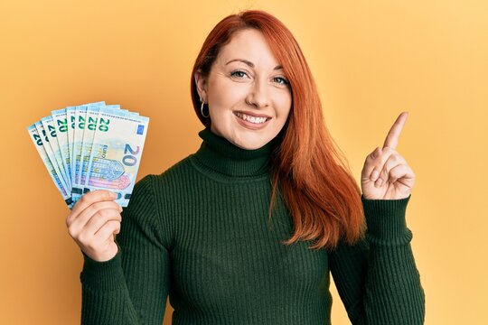 Beautiful redhead woman holding bunch of 20 euro banknotes smiling happy pointing with hand and finger to the side