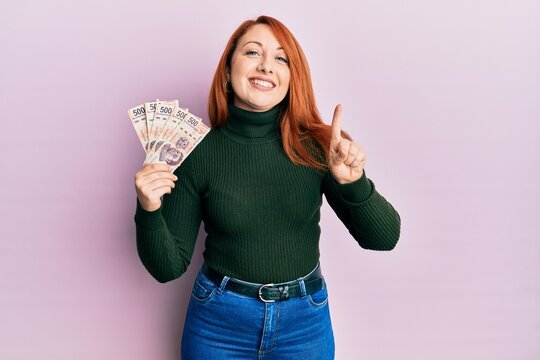Beautiful redhead woman holding 500 mexican pesos banknotes smiling with an idea or question pointing finger with happy face, number one
