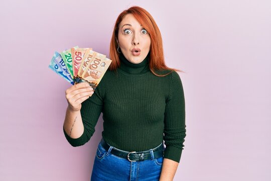 Beautiful redhead woman holding canadian dollars scared and amazed with open mouth for surprise, disbelief face