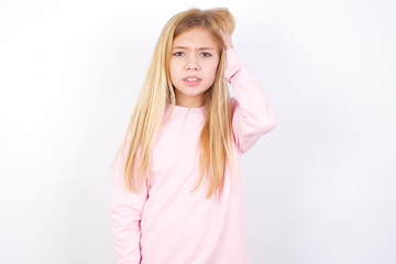 Portrait of confused beautiful caucasian little girl wearing pink hoodie over white wall holding hand on hair and frowning, panicking, losing memory. Worried and anxious can not remember anything.