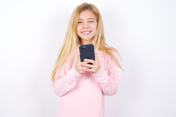 Pleased beautiful caucasian little girl wearing pink hoodie over white background using self phone and looking and winking at the camera. Flirt and coquettish concept.