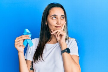 Young hispanic woman holding blue ribbon serious face thinking about question with hand on chin,...