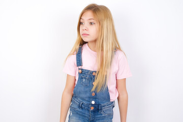 beautiful caucasian little girl wearing jeans overall over white background stares aside with wondered expression has speechless expression. Embarrassed model looks in surprise