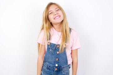 Positive beautiful caucasian little girl wearing jeans overall over white background with overjoyed expression closes eyes and laughs shows white perfect teeth