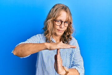 Middle age blonde woman wearing casual clothes and glasses doing time out gesture with hands,...