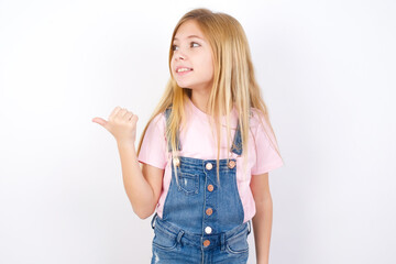 beautiful caucasian little girl wearing jeans overall over white background points away and gives advice demonstrates advertisement