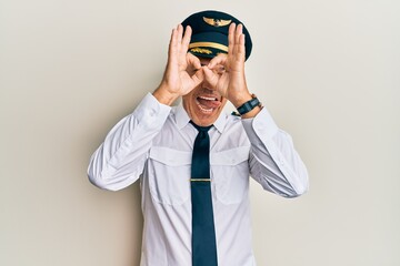 Handsome middle age mature man wearing airplane pilot uniform doing ok gesture like binoculars sticking tongue out, eyes looking through fingers. crazy expression.