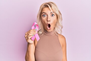 Young blonde girl holding pink cancer ribbon scared and amazed with open mouth for surprise, disbelief face