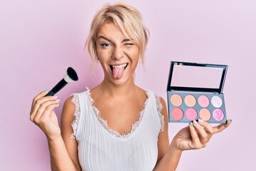 Young blonde girl holding makeup brush and blush sticking tongue out happy with funny expression. - Powered by Adobe