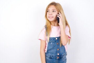 Pleasant looking happy beautiful caucasian little girl wearing jeans overall over white has nice telephone conversation and looks aside, has nice mood and smiles positively while talks via cell phone